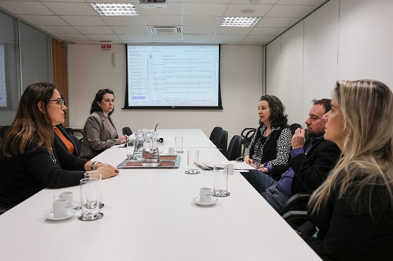 A imagem mostra uma reunião em uma sala de conferências. Há cinco pessoas sentadas ao redor de uma mesa branca retangular. Duas mulheres estão sentadas no lado esquerdo da mesa e um homem e duas mulheres estão no lado direito. Na mesa, há copos de vidro, xícaras de café e uma jarra de água. Ao fundo, há uma tela de projeção com um texto exibido. A sala tem um teto com iluminação embutida e paredes brancas. Há também uma porta de vidro ao fundo, à esquerda, com uma placa de "Saída" ao lado dela.