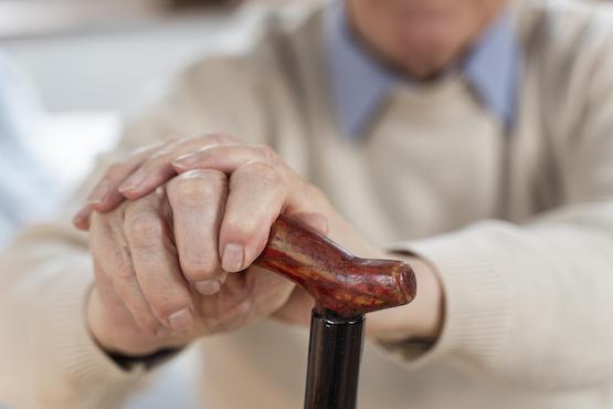 A imagem mostra as mãos de uma pessoa idosa segurando a alça de uma bengala. A pessoa está vestindo um suéter bege e uma camisa azul clara por baixo. A alça da bengala é de madeira com um acabamento polido. A imagem está focada nas mãos e na bengala, com o rosto da pessoa fora de foco ao fundo.