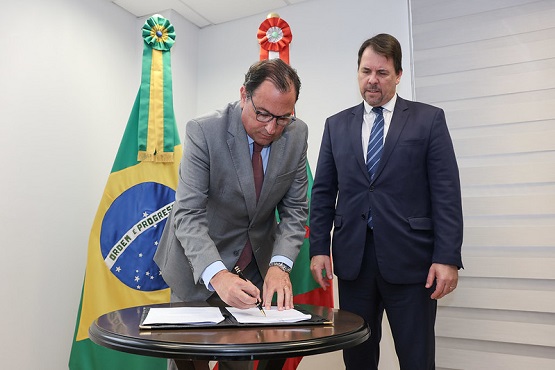 A imagem mostra duas pessoas em um ambiente formal. À esquerda, um homem de terno cinza e gravata vermelha está assinando um documento sobre uma mesa redonda. Ele usa óculos e parece concentrado na assinatura. À direita, outro homem de terno azul escuro e gravata azul está de pé, observando a assinatura. Ao fundo, há duas bandeiras: a bandeira do Brasil à esquerda e a de Santa Catarina à direita. Ambas as bandeiras têm laços decorativos no topo. O ambiente parece ser um escritório ou uma sala de reuniões.