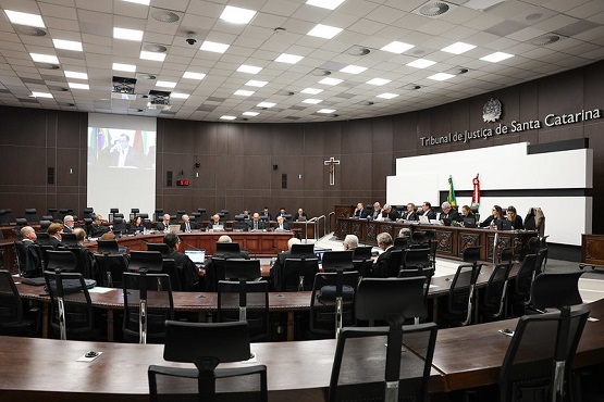 A imagem mostra uma sala de tribunal moderna e espaçosa, com várias pessoas sentadas em cadeiras ao redor de uma grande mesa em formato de "U". Na parede ao fundo, há um crucifixo e as palavras "Tribunal de Justiça de Santa Catarina". Há também duas bandeiras, uma do Brasil e outra do estado de Santa Catarina. No canto superior esquerdo, há uma tela de projeção mostrando a imagem de uma pessoa falando. A sala é bem iluminada, com um teto repleto de luminárias embutidas.
