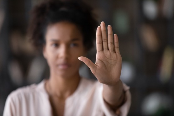 A imagem mostra uma mulher de pele morena e cabelo cacheado, desfocada ao fundo, com a mão direita estendida em direção à câmera, em foco. A palma da mão está aberta, como se estivesse fazendo um gesto de "pare". Ela está usando uma blusa clara e o fundo é desfocado, mas parece ser um ambiente interno com prateleiras e objetos.