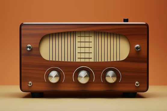 A imagem mostra um rádio antigo de madeira com um design retrô. Ele tem uma frente retangular com bordas arredondadas. No centro, há uma grande tela oval com uma grade de linhas verticais e horizontais, que provavelmente serve para mostrar as frequências das estações de rádio. Abaixo da tela, há três botões redondos de metal, alinhados horizontalmente, que provavelmente são usados para ajustar o volume, a sintonia e outras funções. O fundo da imagem é de cor laranja, e a superfície onde o rádio está apoiado é de cor bege.