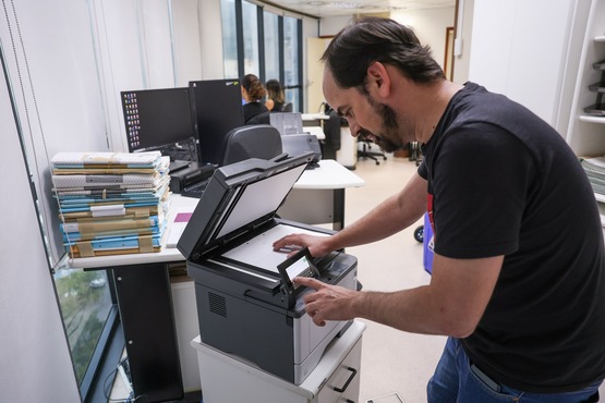 A imagem mostra um homem usando uma impressora multifuncional em um escritório. Ele está de pé ao lado da impressora, com a tampa aberta, colocando ou ajustando um documento no scanner. O homem tem cabelo curto e barba, e está vestindo uma camiseta preta e jeans. Ao fundo, há várias mesas de trabalho com computadores e pilhas de documentos. Algumas pessoas estão sentadas trabalhando nos computadores. O ambiente é bem iluminado, com janelas grandes à esquerda, permitindo a entrada de luz natural.