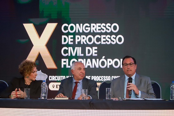 A imagem mostra três pessoas sentadas em uma mesa de conferência. Ao fundo, há um grande painel com o texto "X Congresso de Processo Civil de Florianópolis" e "Circuito Brasileiro de Processo Civil". As três pessoas parecem ser palestrantes ou participantes do evento. A pessoa à esquerda é uma mulher de cabelos cacheados e loiros, vestindo um blazer preto. No centro, há um homem calvo com cabelos grisalhos nas laterais, vestindo um terno cinza e gravata vermelha. À direita, há um homem de óculos, cabelo escuro, vestindo um terno cinza e gravata marrom, que está falando ao microfone. Na mesa, há copos de água e garrafas.