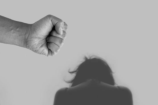 A imagem mostra um punho fechado em destaque à esquerda, sugerindo um gesto de agressão. Ao fundo, há uma pessoa de costas, com a cabeça abaixada e os ombros curvados, aparentando estar em uma posição de tristeza ou medo. A imagem é em preto e branco, o que intensifica o tom dramático e emocional da cena.