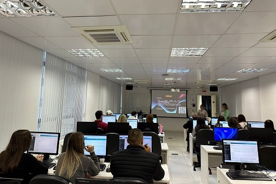A imagem mostra uma sala de aula ou um laboratório de informática. Há várias pessoas sentadas em frente a computadores, olhando para as telas. A sala é bem iluminada, com luzes fluorescentes no teto. Na frente da sala, há uma tela de projeção grande onde parece estar sendo exibida uma apresentação. Algumas pessoas estão de pé, possivelmente o instrutor ou palestrante, e outras estão sentadas, prestando atenção. As paredes são brancas e há um ar-condicionado instalado na parede lateral.
