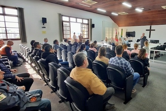 A imagem mostra uma sala de conferências com várias pessoas sentadas em cadeiras, prestando atenção a uma apresentação ou reunião que está acontecendo na frente da sala. O ambiente é iluminado pela luz natural que entra pelas janelas à esquerda. A frente da sala possui uma mesa com várias pessoas sentadas, incluindo uma pessoa de pé que parece estar falando para o público. Há bandeiras ao fundo, indicando possivelmente que a reunião pode ser de caráter oficial ou governamental. O teto da sala é de madeira, e há uma cruz na parede ao fundo, sugerindo que a sala pode estar em uma instituição religiosa ou governamental com afiliação religiosa.