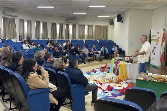 A imagem mostra uma sala de conferências ou auditório com várias pessoas sentadas em cadeiras azuis, dispostas em fileiras. No centro da imagem, há uma mesa coberta com várias toalhas de mesa coloridas e alguns objetos sobre ela. Um homem de camiseta branca está em pé ao lado da mesa, aparentemente falando para o público. As pessoas na audiência parecem estar prestando atenção ao que ele está dizendo.