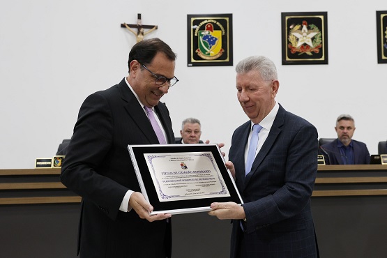 Foto horizontal: Francisco Oliveira Neto recebe homenagem com placa em sessão solene, autoridades ao fundo, ambiente institucional.