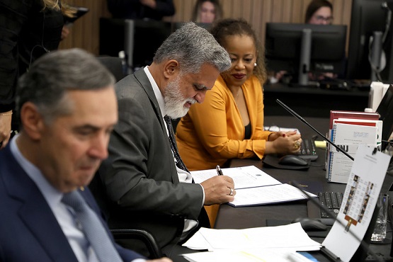 A imagem mostra uma reunião ou conferência com várias pessoas sentadas em uma mesa. A foto está inclinada para a esquerda. Na frente, há um homem de terno cinza e gravata azul claro, com cabelo grisalho, olhando para baixo. Ao lado dele, há outro homem de barba branca e cabelo grisalho, vestindo uma camisa laranja. Ambos estão concentrados em documentos à sua frente. No fundo, há uma mulher de pele clara e cabelo cacheado, também olhando para a mesa. Há papéis, cadernos e microfones sobre a mesa.