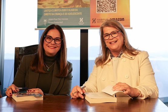 A imagem mostra duas mulheres sentadas lado a lado em uma mesa, ambas sorrindo para a câmera. A mulher à esquerda está usando um casaco marrom escuro e óculos, enquanto a mulher à direita está usando um casaco bege claro e também usa óculos. Ambas estão segurando papéis ou documentos. Ao fundo, há um banner com texto e um QR code, mas o conteúdo exato do texto não é legível na imagem.