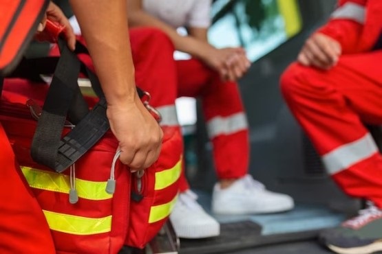 A imagem mostra três pessoas vestindo uniformes vermelhos com faixas refletivas amarelas, típicos dos bombeiros. Uma das pessoas está segurando uma bolsa vermelha com faixas refletivas amarelas, que parece ser um kit de primeiros socorros. As outras duas pessoas estão sentadas, e uma delas está usando tênis brancos. A cena parece estar dentro de uma ambulância ou veículo de emergência.