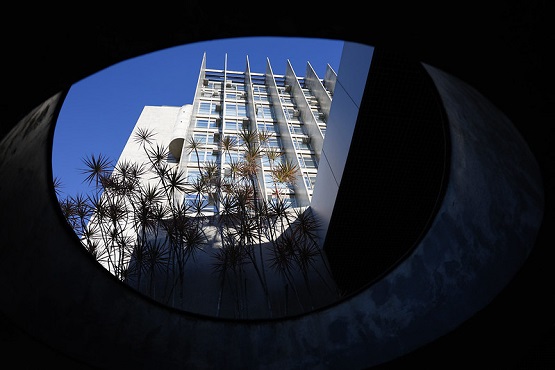 A imagem mostra a vista de um prédio moderno através de uma abertura circular no teto de uma estrutura. O prédio tem uma fachada branca com várias janelas e elementos verticais que parecem ser colunas ou ripas. No centro da imagem, há algumas plantas altas com folhas finas e pontiagudas. O céu ao fundo está azul e sem nuvens, proporcionando um contraste nítido com a estrutura branca do prédio.