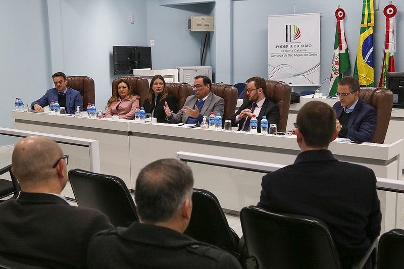 A imagem mostra uma reunião formal em uma sala de conferências. Há seis pessoas sentadas em uma mesa comprida, todas vestidas formalmente. Da direita da foto para a esquerda, há três homens, duas mulheres e mais um homem. Eles estão sentados em cadeiras de couro marrom. Na frente deles, há garrafas de água e copos. Atrás da mesa, na parede, há um banner com o logotipo do Poder Judiciário de Santa Catarina e a inscrição "Comarca de São Miguel do Oeste". À direita do banner, há três bandeiras: a do Brasil, a de Santa Catarina e uma terceira bandeira que não é claramente visível. Na frente da mesa, há algumas pessoas sentadas, de costas para a câmera, assistindo à reunião. A sala tem paredes claras e uma atmosfera formal.