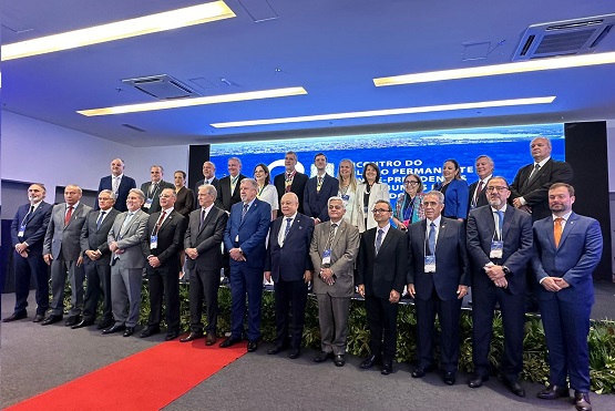 A imagem mostra um grupo de aproximadamente 27 homens e mulheres posando para uma foto em um evento formal. Eles estão em um palco, organizados em duas fileiras: uma fileira de pessoas em pé na parte de trás e outra fileira de pessoas em pé na frente. Todos estão vestidos formalmente, com ternos e gravatas, e algumas mulheres estão usando vestidos ou ternos femininos. Ao fundo, há um grande painel azul com texto branco, mas o texto não está totalmente legível na imagem. O palco tem uma decoração simples com algumas plantas na frente. O ambiente parece ser um auditório ou uma sala de conferências, com iluminação de teto moderna. Há também um tapete vermelho no chão, que leva até o palco.