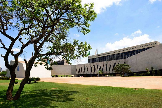 A imagem mostra um edifício moderno com arquitetura de linhas retas e formas geométricas, localizado em um espaço aberto com bastante área verde. À esquerda, há uma árvore com galhos espalhados e folhas verdes. O céu está claro com algumas nuvens brancas. O edifício tem uma fachada de concreto com janelas na parte superior e uma série de elementos verticais decorativos na parte inferior. Há também uma bandeira do Brasil visível no topo do edifício. A área em frente ao edifício é pavimentada, com algumas árvores e arbustos espalhados.