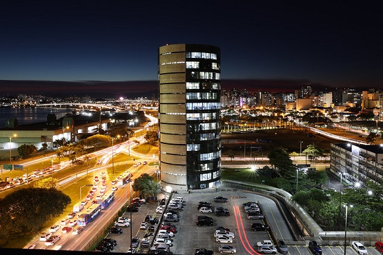 A imagem mostra uma paisagem urbana à noite. No centro, há um edifício alto e cilíndrico com várias janelas iluminadas. Ao redor do edifício, há ruas movimentadas com muitos carros, cujas luzes formam trilhas devido à longa exposição da foto. À esquerda, há uma área com árvores e um corpo d'água ao fundo. À direita, há mais edifícios e áreas urbanas iluminadas. O céu está escuro, indicando que é noite.
