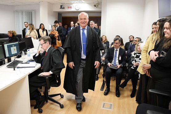 A imagem mostra um ambiente interno, provavelmente uma sala de tribunal ou uma sala de conferências. No centro da imagem, há um homem mais velho, de cabelos brancos, usando uma toga preta e uma gravata azul, caminhando em direção à câmera com um sorriso no rosto. Ele parece ser uma figura de autoridade. À esquerda, há um homem sentado em uma cadeira de escritório, também usando uma toga preta, olhando para um monitor de computador. Atrás dele, há mais pessoas sentadas e em pé, algumas conversando entre si. À direita, há um grupo de pessoas sentadas, algumas sorrindo e olhando para o homem que está caminhando. O ambiente é bem iluminado, com paredes brancas e piso de madeira clara. Há uma sensação de formalidade e seriedade no local, mas também um clima de leveza e descontração, possivelmente devido ao sorriso do homem que está caminhando.