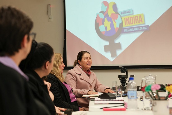 A imagem mostra uma reunião em uma sala de conferências. Há várias pessoas sentadas ao redor de uma mesa, prestando atenção em uma mulher que está falando. Ela está vestindo um casaco rosa claro e parece ser a principal oradora. Na mesa, há diversos itens como garrafas de água, copos, cadernos e canetas. Ao fundo, há uma tela de projeção com o logotipo do "Programa Indira - Pelas Mulheres do PJSC". O logotipo inclui um símbolo feminino com várias silhuetas coloridas de pessoas dentro dele. A sala parece bem iluminada e as pessoas estão concentradas na apresentação.