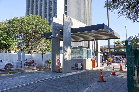 A imagem mostra uma entrada de um edifício com uma estrutura de cobertura moderna. Há cones de trânsito laranja e brancos no chão, delimitando uma área de passagem. Ao fundo, é possível ver árvores e um edifício alto.