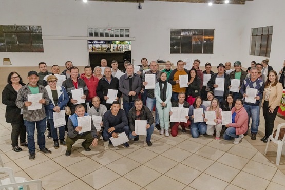 A imagem mostra um grupo grande de pessoas posando para uma foto em um ambiente interno. A maioria das pessoas está segurando certificados ou diplomas, sugerindo que acabaram de participar de uma cerimônia de formatura ou entrega de certificados. Eles estão organizados em várias fileiras, com algumas pessoas agachadas na frente e outras em pé atrás. O ambiente parece ser uma sala com piso de cerâmica e paredes brancas. Há algumas janelas visíveis ao fundo.