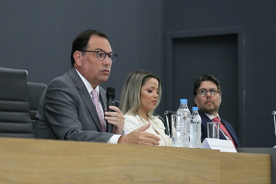 A imagem mostra três pessoas sentadas lado a lado em uma mesa, aparentemente participando de uma conferência ou reunião. A pessoa à esquerda está falando ao microfone e usa óculos, um terno cinza e uma gravata rosa. Ao centro, há uma mulher com cabelo loiro, vestindo uma blusa branca. À direita, um homem de óculos, barba e cabelo castanho, usando um terno escuro e gravata vermelha. Na mesa, há garrafas de água e copos. O fundo é de cor escura.