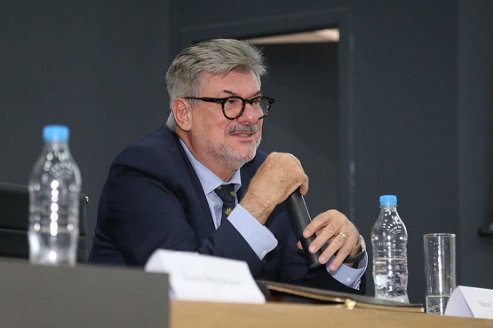 A imagem mostra um homem de cabelos grisalhos e barba, usando óculos, terno e gravata, segurando um microfone enquanto parece estar falando. Ele está sentado à mesa, que tem uma garrafa de água e um copo. Ao fundo, há uma parede escura. A imagem está inclinada para a esquerda.