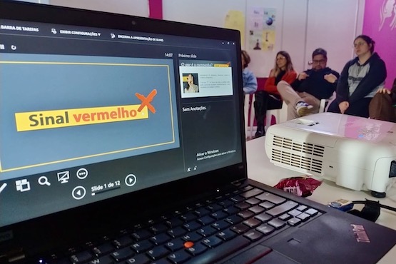 A imagem mostra uma apresentação de slides em um laptop, com o slide atual exibindo o título "Sinal vermelho" em destaque. O slide está sendo projetado em uma sala onde algumas pessoas estão sentadas ao fundo, prestando atenção. O projetor está posicionado sobre uma mesa ao lado do laptop. O ambiente é iluminado e as paredes têm algumas decorações e cartazes.