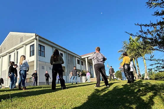 A imagem mostra um grupo de pessoas em um gramado em frente a um prédio de dois andares. O céu está azul e ensolarado. As pessoas estão espalhadas pelo gramado em círculo, sendo que há um homem no centro. O prédio ao fundo é de cor clara, com janelas grandes e uma estrutura que parece ser de concreto. Há também algumas árvores, incluindo uma palmeira, à direita da imagem. As sombras no gramado indicam que o sol está brilhando intensamente. Parece ser um dia agradável e as pessoas estão vestidas de forma casual.
