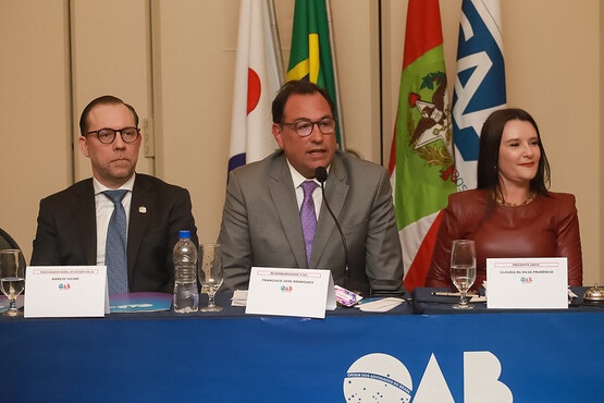A imagem mostra três pessoas sentadas atrás de uma mesa coberta com uma toalha azul que tem o logotipo da OAB (Ordem dos Advogados do Brasil). No centro, há um homem de terno cinza e gravata roxa, falando em um microfone. À sua esquerda, está um homem de óculos, terno preto e gravata azul. À direita, uma mulher com cabelo castanho liso, vestindo uma blusa de couro marrom. Atrás deles, há bandeiras, incluindo a do Brasil e de Santa Catarina. Na mesa, há placas com os nomes das pessoas, copos de água e uma garrafa de água. Parece ser um evento formal ou uma conferência.