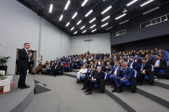A imagem mostra um auditório cheio de pessoas sentadas em cadeiras azuis, todas voltadas para um palco onde um homem de terno está em pé, aparentemente falando ou apresentando algo. O auditório é grande, com um teto alto e várias luzes no teto. As pessoas parecem estar prestando atenção ao que está sendo dito. O ambiente é formal, sugerindo que pode ser uma conferência, palestra ou evento corporativo.
