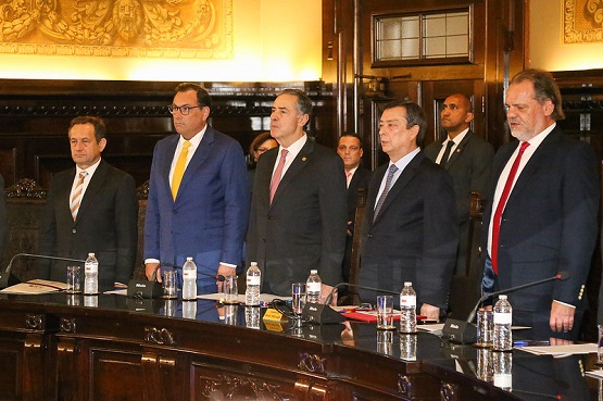 A imagem mostra um grupo de seis homens em pé, lado a lado, em um ambiente formal que parece ser uma sala de reuniões ou um tribunal. Todos estão vestidos com ternos escuros e gravatas de cores variadas. Eles estão posicionados atrás de uma mesa comprida, que tem microfones e garrafas de água sobre ela. Ao fundo, há detalhes decorativos nas paredes, incluindo uma bandeira do Brasil visível à esquerda. A cena sugere um evento oficial ou uma cerimônia.