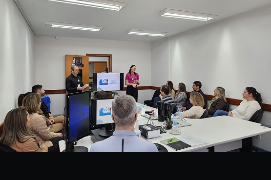 A imagem mostra uma sala de conferência com várias pessoas. Na parte superior, há um grupo de pessoas sentadas ao redor de uma mesa branca, olhando para a frente, onde há uma mulher em pé, vestindo uma blusa rosa, que parece estar apresentando algo. Atrás dela, há duas telas exibindo o mesmo slide, que parece ser parte de uma apresentação. Na parte inferior da imagem, há um homem em pé, sorrindo, vestindo uma jaqueta preta e calça jeans, com um crachá pendurado no pescoço. Ele está posicionado ao lado de uma mesa com garrafas de água. A sala tem paredes brancas e iluminação fluorescente no teto.