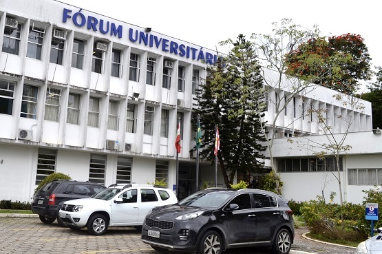 A imagem mostra a fachada de um prédio de três andares identificado como "Fórum Universitário". O edifício é de cor branca e possui várias janelas retangulares. Na frente do prédio, há uma área de estacionamento onde estão estacionados alguns carros, incluindo um SUV preto e um carro branco. Há também algumas árvores e arbustos ao redor, e bandeiras hasteadas próximas à entrada do edifício. O céu está nublado, sugerindo um dia sem sol.