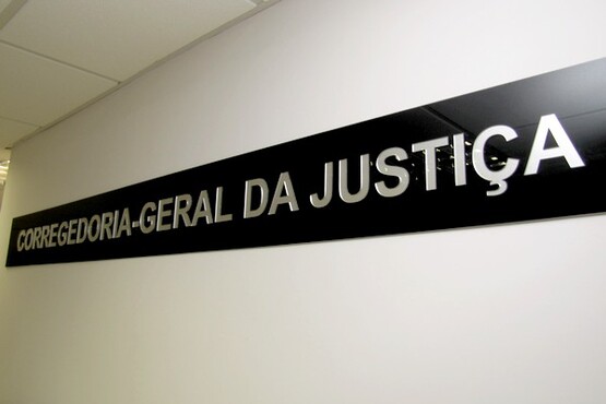 A imagem mostra uma placa preta com letras prateadas em alto relevo, fixada em uma parede clara. Na placa está escrito "CORREGEDORIA-GERAL DA JUSTIÇA". A placa parece estar em um ambiente interno, possivelmente em um prédio governamental ou judiciário. O teto é visível na parte superior da imagem, com iluminação embutida.
