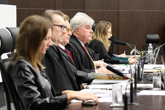 A imagem mostra uma mesa de tribunal com cinco pessoas sentadas lado a lado. Três delas são homens e duas são mulheres. Todos estão vestindo togas pretas. Eles estão sentados em cadeiras pretas e há microfones à frente de cada um. A mesa está coberta com papéis, copos de água e garrafas. A parede ao fundo é de madeira escura, conferindo um ar formal ao ambiente. As pessoas parecem estar concentradas, possivelmente participando de uma audiência ou julgamento.