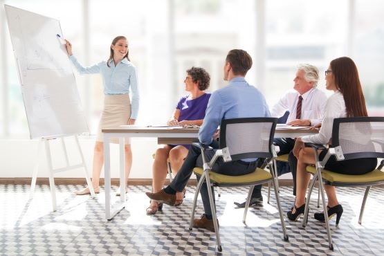 A imagem mostra uma reunião de negócios em uma sala de conferências. Há cinco pessoas na cena. Uma mulher está em pé ao lado de um quadro branco, apontando para ele enquanto fala, possivelmente apresentando ou explicando algo. Ela está vestida com uma camisa azul clara e uma saia bege. Sentados ao redor de uma mesa, há quatro pessoas: duas mulheres e dois homens. Uma das mulheres está usando uma blusa roxa e óculos, enquanto a outra está de camisa branca e calça preta. Um dos homens está de camisa azul clara e o outro de camisa branca e gravata. Todos parecem estar prestando atenção na apresentação. A sala é bem iluminada, com janelas grandes ao fundo, e o chão tem um padrão geométrico.