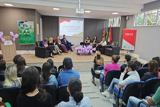 A imagem mostra um auditório onde está acontecendo um evento ou palestra. No palco, há sete pessoas sentadas em poltronas pretas, participando de uma discussão. Atrás delas, há uma tela de projeção com um slide exibindo o título que não é legível. No palco, também há várias decorações em formato de laço na cor lilás, que é frequentemente associada a campanhas de conscientização sobre a violência contra a mulher. À esquerda, há um banner verde com imagens de pessoas e à direita, um banner vermelho com a palavra "DIREITO". O público está sentado em cadeiras, assistindo à apresentação. As paredes do auditório são cinzas e há janelas no lado direito, permitindo a entrada de luz natural.