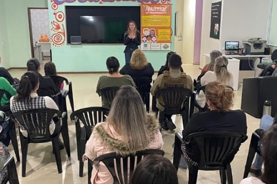 A imagem mostra uma sala onde várias pessoas estão sentadas em cadeiras de plástico preto, voltadas para a frente, onde uma mulher está de pé, aparentemente dando uma palestra ou apresentação. A mulher está em frente a uma grande televisão ou tela e uma lousa de giz, que está desligada ou mostrando uma imagem escura. Atrás dela, há um painel colorido com textos e imagens, mas os detalhes não são legíveis. As pessoas na audiência estão prestando atenção na apresentação. A sala parece ser um espaço comunitário ou de eventos, com paredes claras e decoração simples.