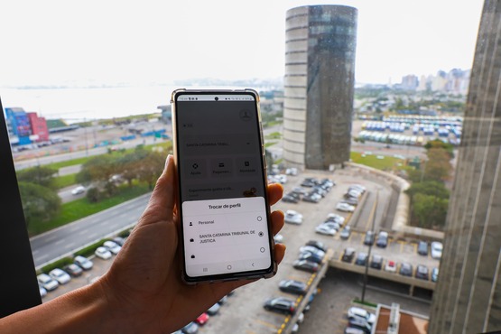 Imagem mostra um celular com um aplicativo aberto, em segundo plano, ao fundo, mostra uma janela com vista para uma cidade.