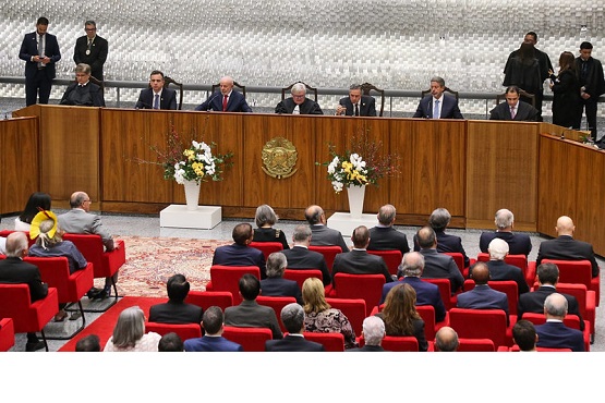 Foto horizontal: sessão solene em plenário do Judiciário, autoridades à mesa e público em cadeiras vermelhas, ambiente institucional.