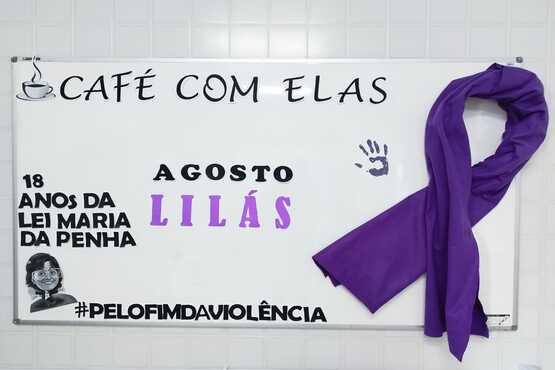 A imagem mostra um quadro branco com várias inscrições e um laço roxo pendurado no lado direito. No topo do quadro está escrito "CAFÉ COM ELAS" com um desenho de uma xícara de café ao lado esquerdo. Abaixo, à esquerda, está escrito "18 ANOS DA LEI MARIA DA PENHA" com uma ilustração do rosto de uma mulher. No centro, está escrito "AGOSTO LILÁS" com a palavra "LILÁS" em destaque na cor lilás. Há também uma marca de mão pintada em roxo. Na parte inferior, está a hashtag "#PELOFIMDAVIOLÊNCIA". A imagem parece estar promovendo a conscientização sobre a violência contra a mulher, destacando o "Agosto Lilás", uma campanha de combate à violência doméstica.