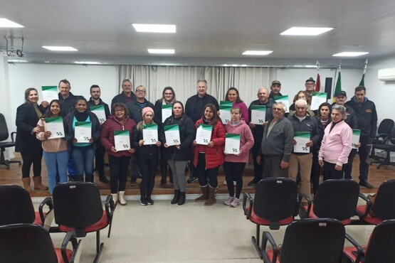 A imagem mostra um grupo de pessoas posando para uma foto em uma sala de conferências ou auditório. Há cerca de 25 pessoas, a maioria segurando certificados ou pastas com uma capa verde e branca. Eles estão em pé em duas fileiras, com algumas pessoas na frente e outras atrás. A sala tem cadeiras vazias na frente do grupo e cortinas brancas ao fundo. Há também bandeiras no canto direito da imagem. As pessoas estão vestidas de forma casual, com roupas de frio, como casacos e botas. Parece ser uma cerimônia de entrega de certificados, diplomas ou documentos.
