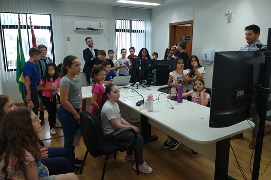 A imagem mostra um ambiente educacional onde há um grupo de crianças e alguns adultos. As crianças estão reunidas em torno de uma mesa grande que possui vários computadores e monitores. Algumas crianças estão sentadas, enquanto outras estão em pé, olhando para os monitores. Há dois adultos na sala, um deles está em pé ao lado da mesa, aparentemente explicando algo para as crianças, e o outro está ao fundo, próximo à janela, observando a cena. A sala é bem iluminada, com janelas ao fundo e cortinas brancas. As crianças parecem estar atentas e interessadas no que está sendo mostrado nos computadores.
