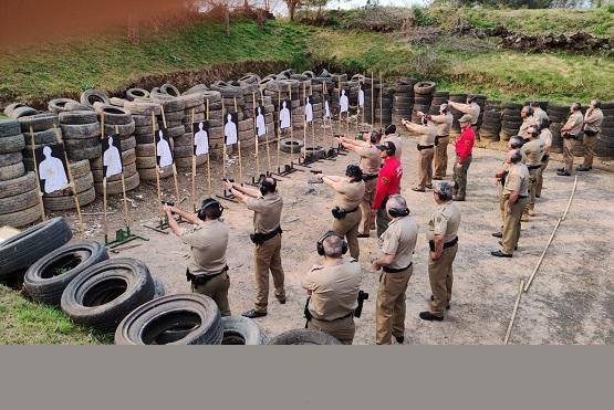 A imagem mostra um grupo de policiais em um campo de treinamento de tiro ao ar livre. Eles estão posicionados em linha, cada um apontando uma arma em direção a alvos de papel que têm a forma de silhuetas humanas. Os alvos estão fixados em suportes de madeira e dispostos em frente a uma parede de pneus empilhados, que provavelmente serve como barreira de segurança. Os policiais estão usando uniformes bege e protetores auriculares. Há também algumas pessoas vestidas de vermelho, que podem ser instrutores ou supervisores. O ambiente ao redor é de terra batida, com vegetação ao fundo.