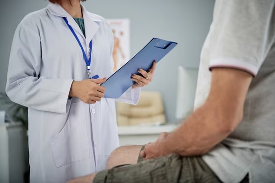 A imagem mostra uma cena em um consultório médico. À esquerda, há uma pessoa vestida com um jaleco branco, segurando uma prancheta azul. Ela parece estar conversando com um paciente, que está parcialmente visível à direita da imagem. O paciente está sentado, vestindo uma camiseta cinza e shorts. O ambiente é claro e parece ser uma sala de consulta médica, com móveis e equipamentos ao fundo.