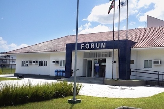 A imagem mostra a fachada de um prédio identificado como "Fórum". O edifício é de um andar, com paredes brancas e um telhado de telhas vermelhas. Na entrada principal, há uma estrutura revestida de azulejos azuis escuros, onde está escrito "FÓRUM" em letras brancas. Em frente ao prédio, há uma área pavimentada e um gramado com algumas plantas. Três mastros com bandeiras estão posicionados à direita da entrada. O céu está claro e azul, com algumas nuvens brancas.