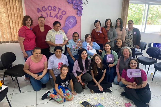 A imagem mostra um grupo de mulheres reunidas em uma sala. Elas estão posando para uma foto em frente a um painel decorativo que diz "Agosto Lilás", uma campanha de conscientização sobre a violência contra a mulher. O painel é decorado com flores roxas e cartazes. As mulheres estão sorrindo e algumas seguram placas roxas com mensagens de apoio à campanha. A sala tem cadeiras dispostas ao redor e uma janela ao fundo, que deixa entrar luz natural. No chão, há um tapete circular com alguns objetos em cima.