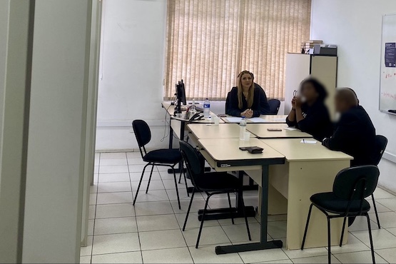 A imagem mostra uma sala de escritório com três pessoas sentadas ao redor de uma mesa de reunião. A sala tem paredes brancas e uma janela com persianas fechadas ao fundo. A mesa é grande e em formato de "L", com um computador e alguns papéis sobre ela. À esquerda, uma mulher loira está sentada de frente para a câmera, enquanto as outras duas pessoas, um homem e uma mulher, estão sentadas de costas para a câmera, com seus rostos desfocados. Há cadeiras de escritório ao redor da mesa e o chão é de azulejos brancos. No canto da sala, há um armário alto. A atmosfera parece ser de uma reunião ou discussão de trabalho.