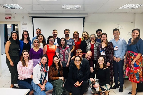 A imagem mostra um grupo de pessoas posando para uma foto em um ambiente interno. Há cerca de 23 pessoas, a maioria mulheres, e elas estão organizadas em três fileiras. Algumas estão sentadas no chão, outras em cadeiras, e algumas em pé. As pessoas estão sorrindo e parecem estar em um ambiente de trabalho ou evento. Ao fundo, há uma tela de projeção e uma parede branca.